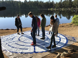 A group of people standing on a portable labyrinth with a scenic view of trees and water in the background.