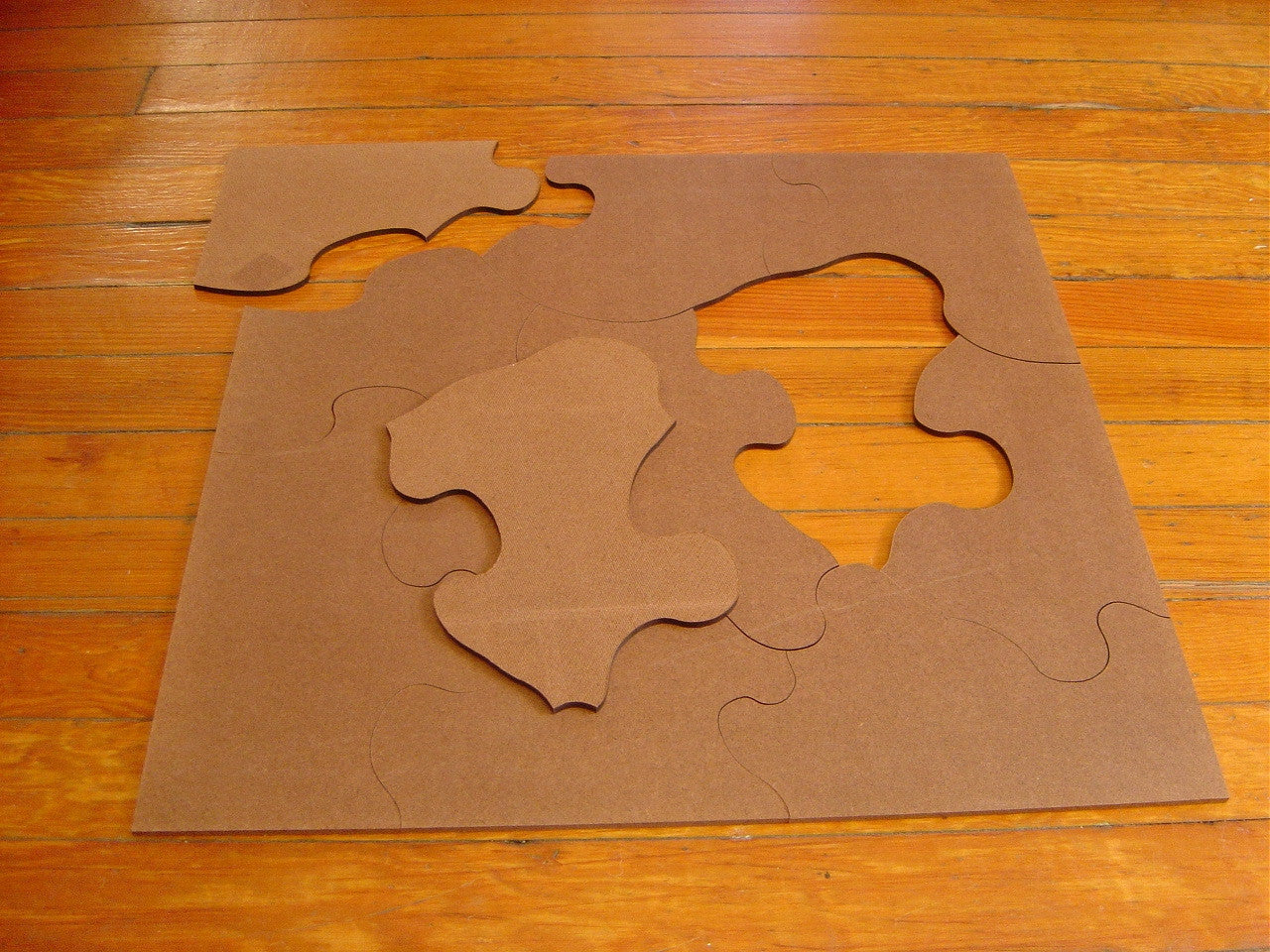 A large, flat, brown cardboard jigsaw puzzle with several pieces arranged on a wooden floor.